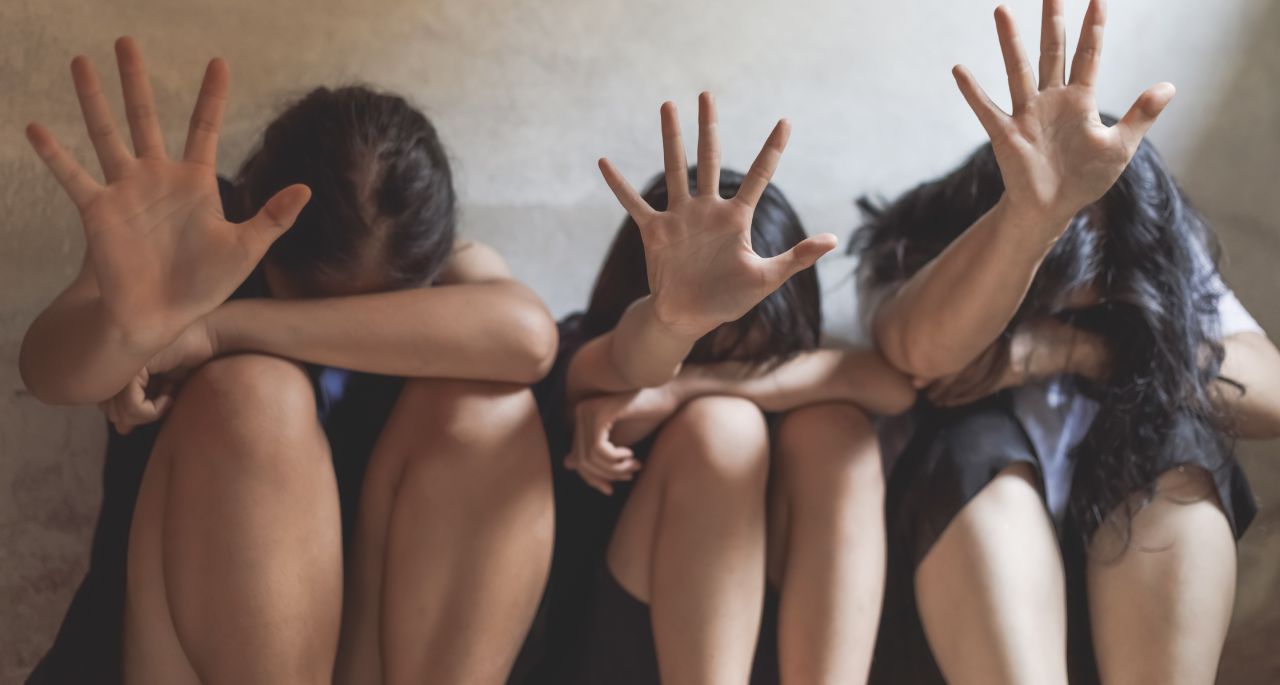 Four people sit against a wall with their heads down and hands raised, palms facing forward, appearing to shield themselves. The scene conveys a sense of fear or distress.