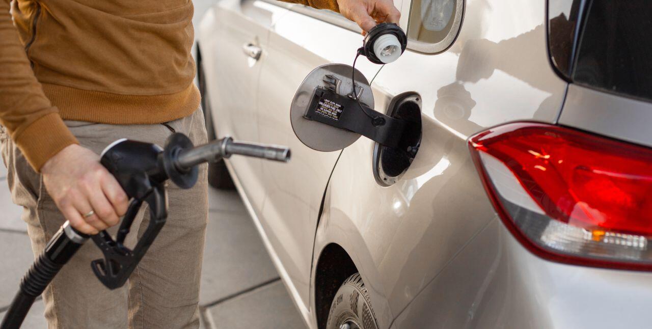 A person refuels a silver car at a gas station, holding a fuel nozzle in one hand and the car's gas cap in the other. The car is parked on a concrete surface. The setting appears to be outdoors, during the daytime.