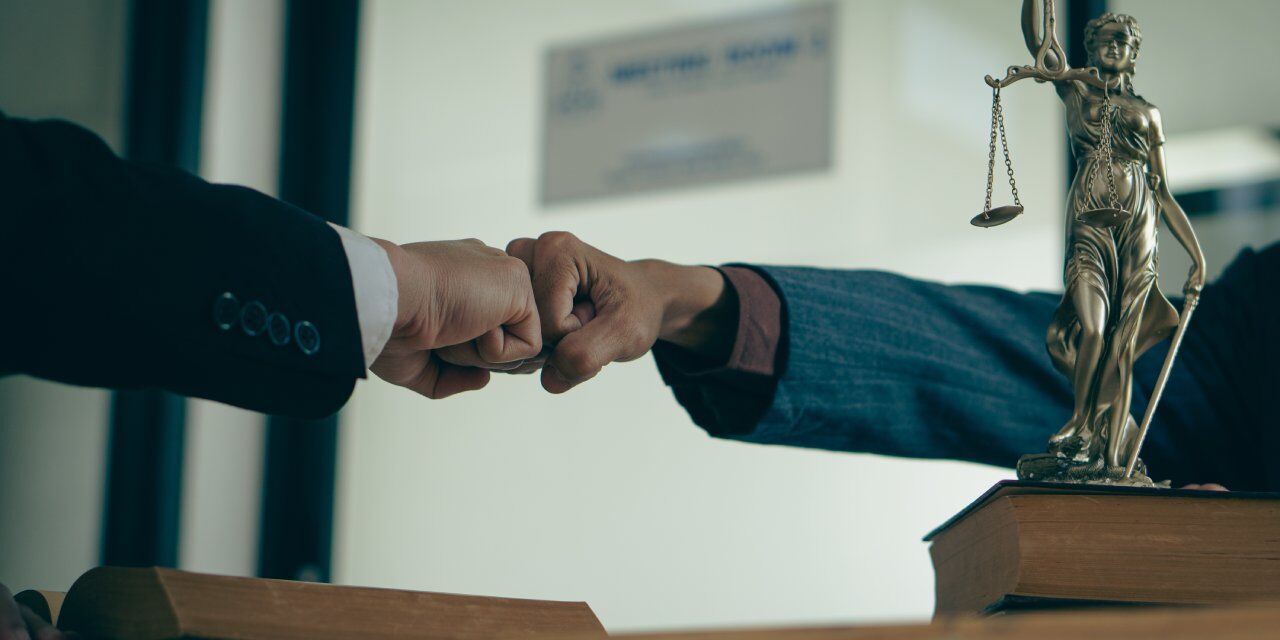 Two people fist-bump over a desk adorned with law books and a statue of Lady Justice, symbolizing legal collaboration or agreement.