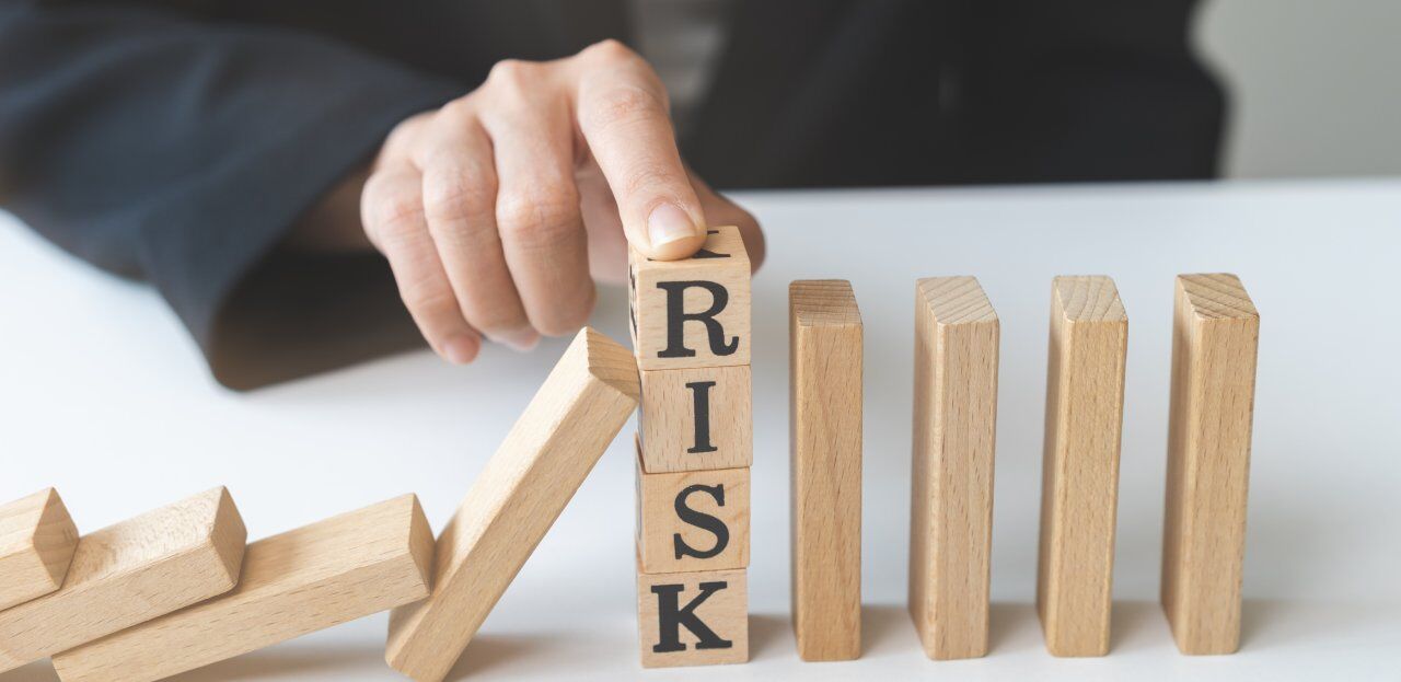 A hand stops toppling wooden dominoes with blocks spelling "RISK." The dominoes on the left are falling, while the hand holds the "RISK" blocks steady, preventing further collapse.