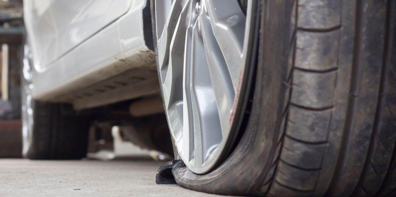 Close-up view of a flat tire on a parked silver car. The tire is deflated, with the sidewall visibly compressed against the pavement, and the wheel rim is prominently featured. The background is slightly blurred.