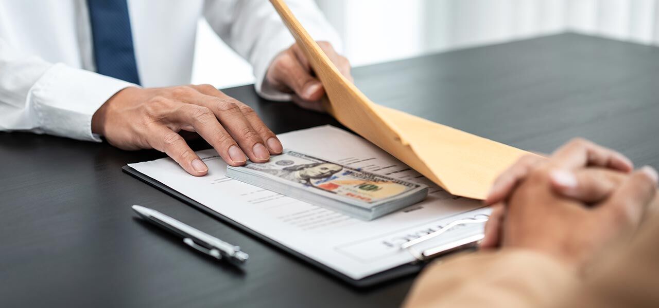 A person hands a brown envelope with a stack of hundred-dollar bills on top of a clipboard containing a document, across a table. A pen lies nearby, and another person with folded hands listens attentively.