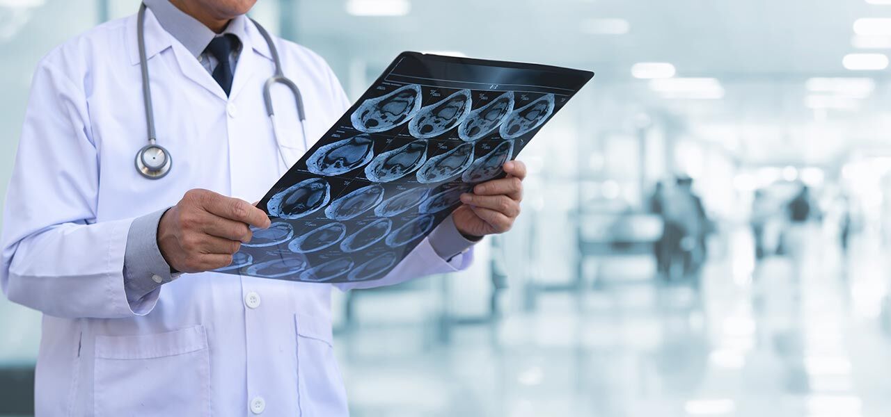 A doctor in a white coat and stethoscope examines a series of X-ray or MRI scans in a bright hospital hallway. The images display various cross-sectional views, and the background is blurred with medical equipment visible.