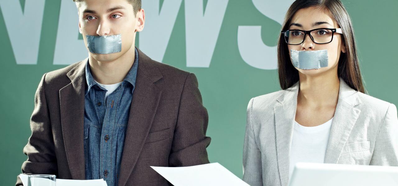 Two people sitting side by side with duct tape over their mouths, holding papers. They're dressed in business attire against a green background with the partial word "NEWS" visible.