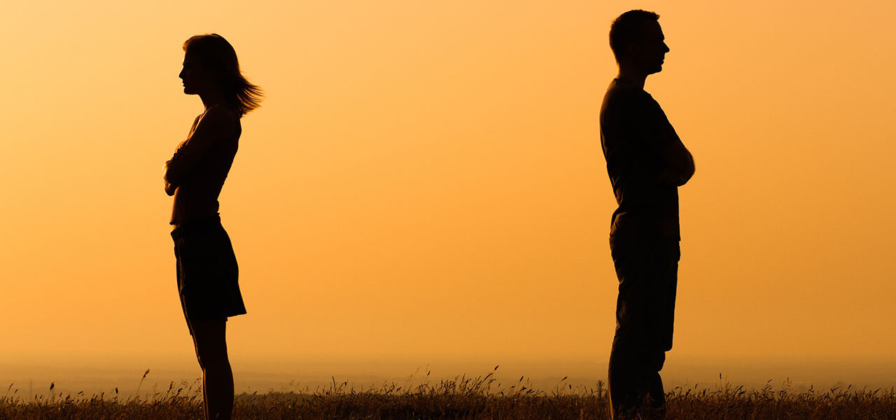 Silhouettes of a man and a woman standing back-to-back with folded arms against a sunset backdrop, suggesting tension or disagreement. The scene conveys a sense of distance or emotional separation.
