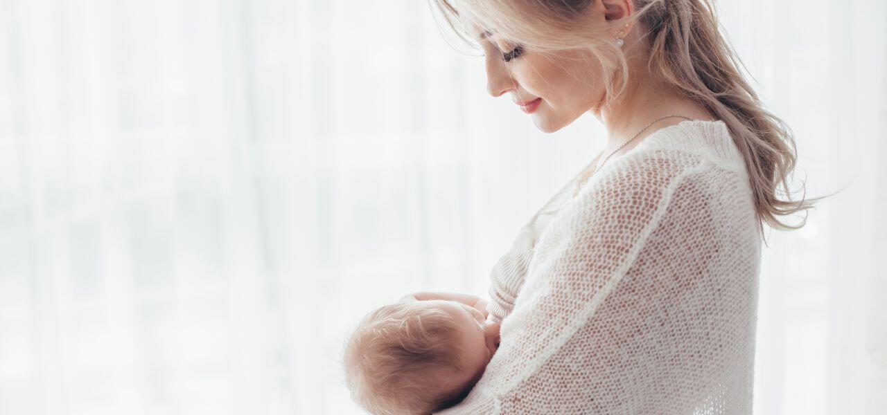 A woman with blonde hair is gently holding and breastfeeding a baby. She is wearing a light knitted sweater, and they are near a softly lit window with sheer curtains. The scene conveys warmth and tenderness.