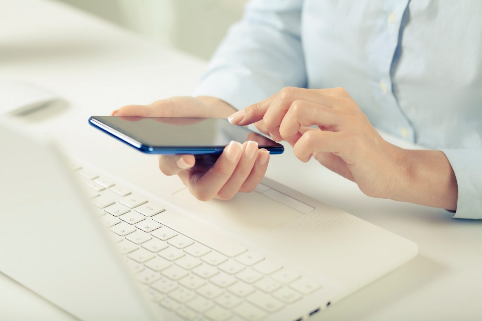 A person in a light blue shirt is using a smartphone in front of an open white laptop. The person is sitting at a desk, and their attention is focused on the smartphone screen.