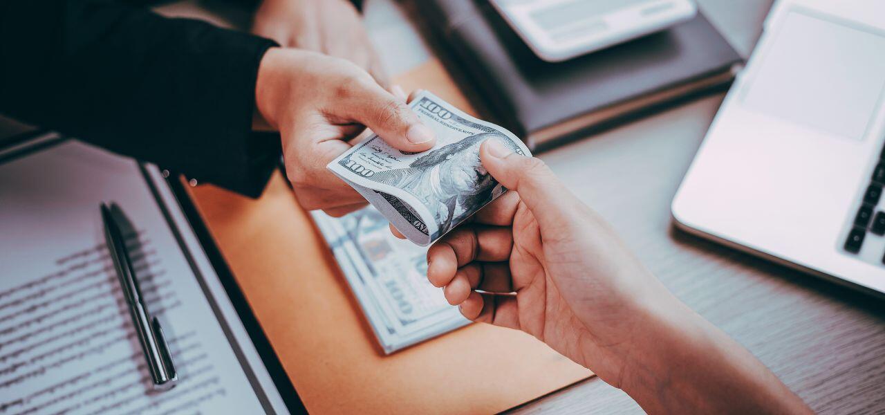 Two hands exchanging a stack of U.S. hundred-dollar bills over a table with documents, a laptop, and a calculator. One hand is giving, and the other is receiving the money.