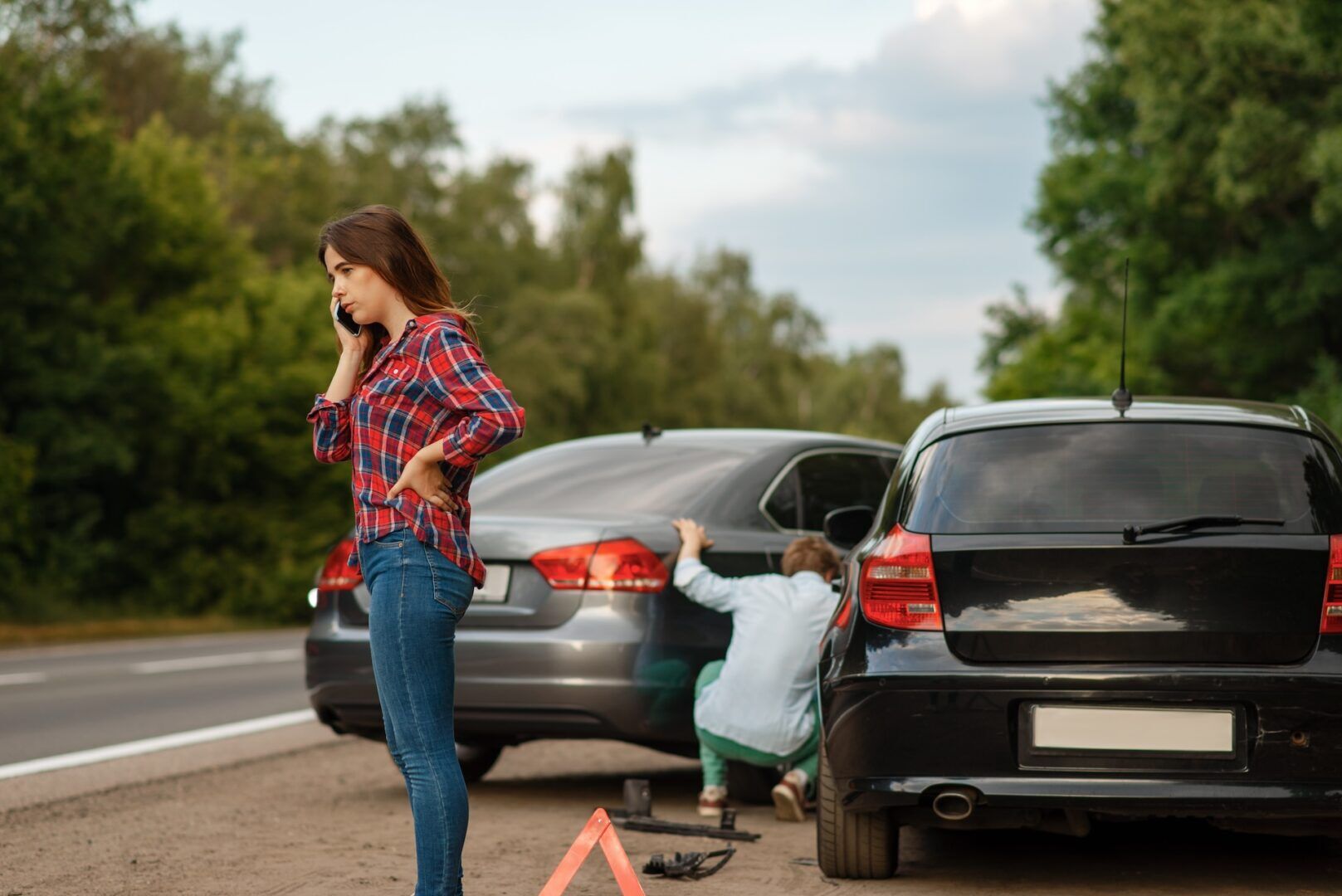 A woman standing by the roadside, talking on her phone, looking concerned. Two cars are parked on the shoulder, with a man kneeling by one car's open trunk. A warning triangle is placed on the road. Trees line the background. A woman standing by the roadside, talking on her phone, looking concerned. Two cars are parked on the shoulder, with a man kneeling by one car's open trunk. A warning triangle is placed on the road. Trees line the background.
