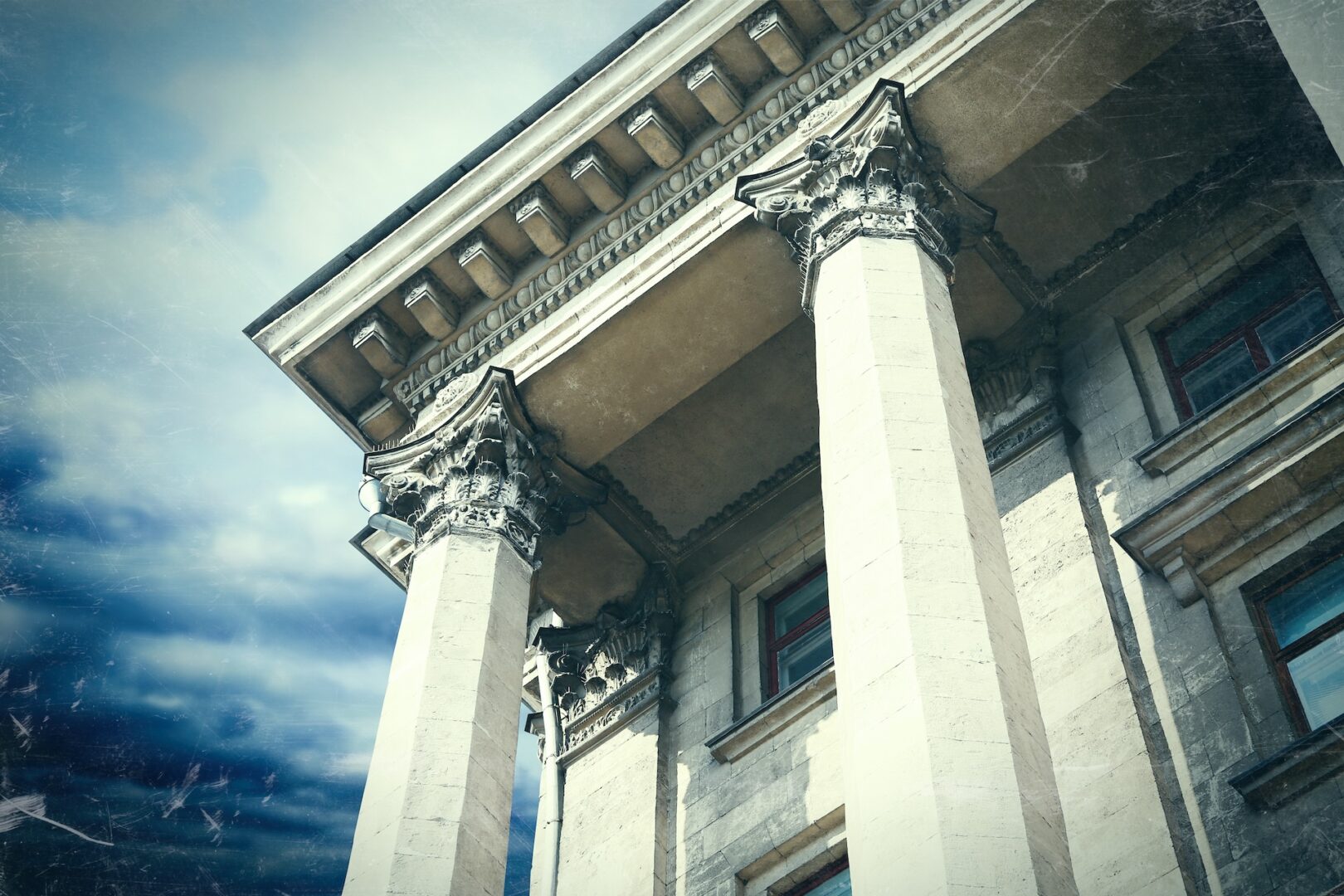 Close-up of an ancient building's facade, featuring three tall, ornate columns with decorative capitals. The structure appears weathered, adding a vintage feel. The sky above is cloudy, enhancing the dramatic atmosphere.