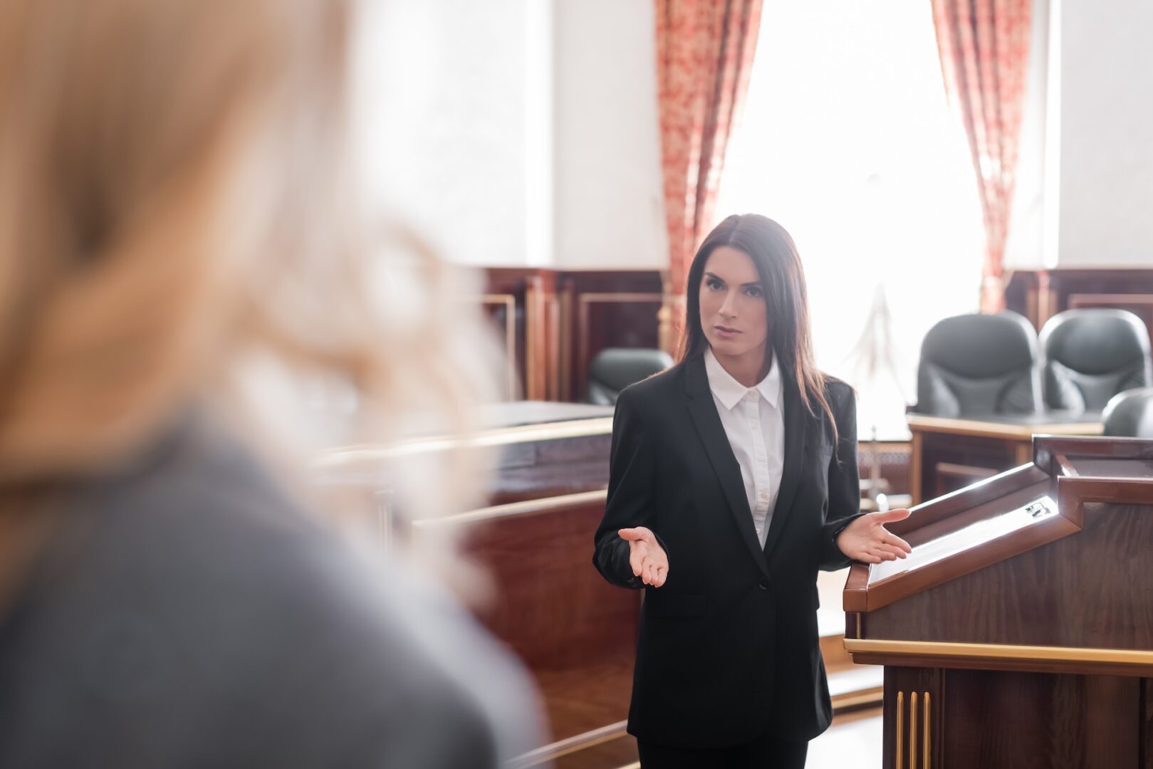 A woman in a suit stands at a podium in a courtroom, gesturing with her hands as if speaking or presenting. The room features wood paneling and red curtains, with empty chairs in the background. Another person is partially visible in the foreground.