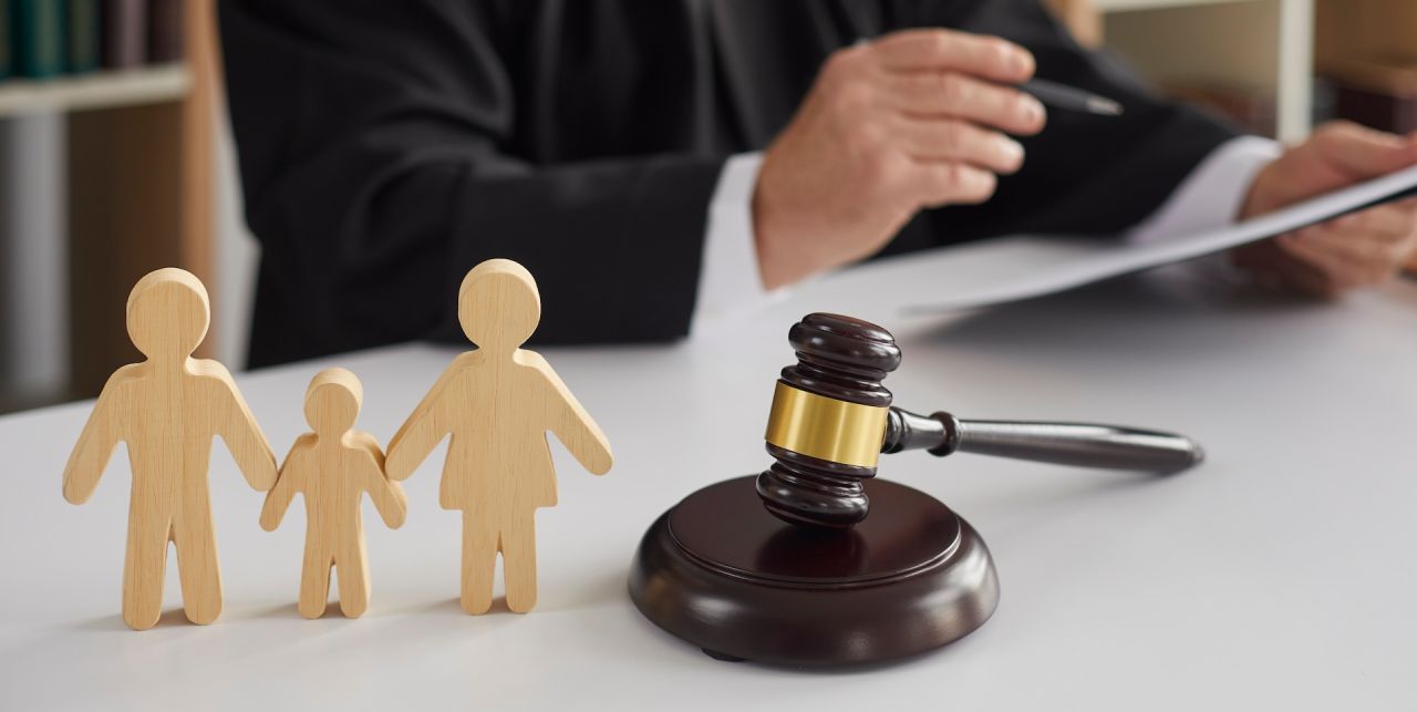 A judge in a black robe holds a pen and paper at a desk. In the foreground, wooden figures of a family and a gavel are positioned on the table. The scene suggests a legal or family court setting.