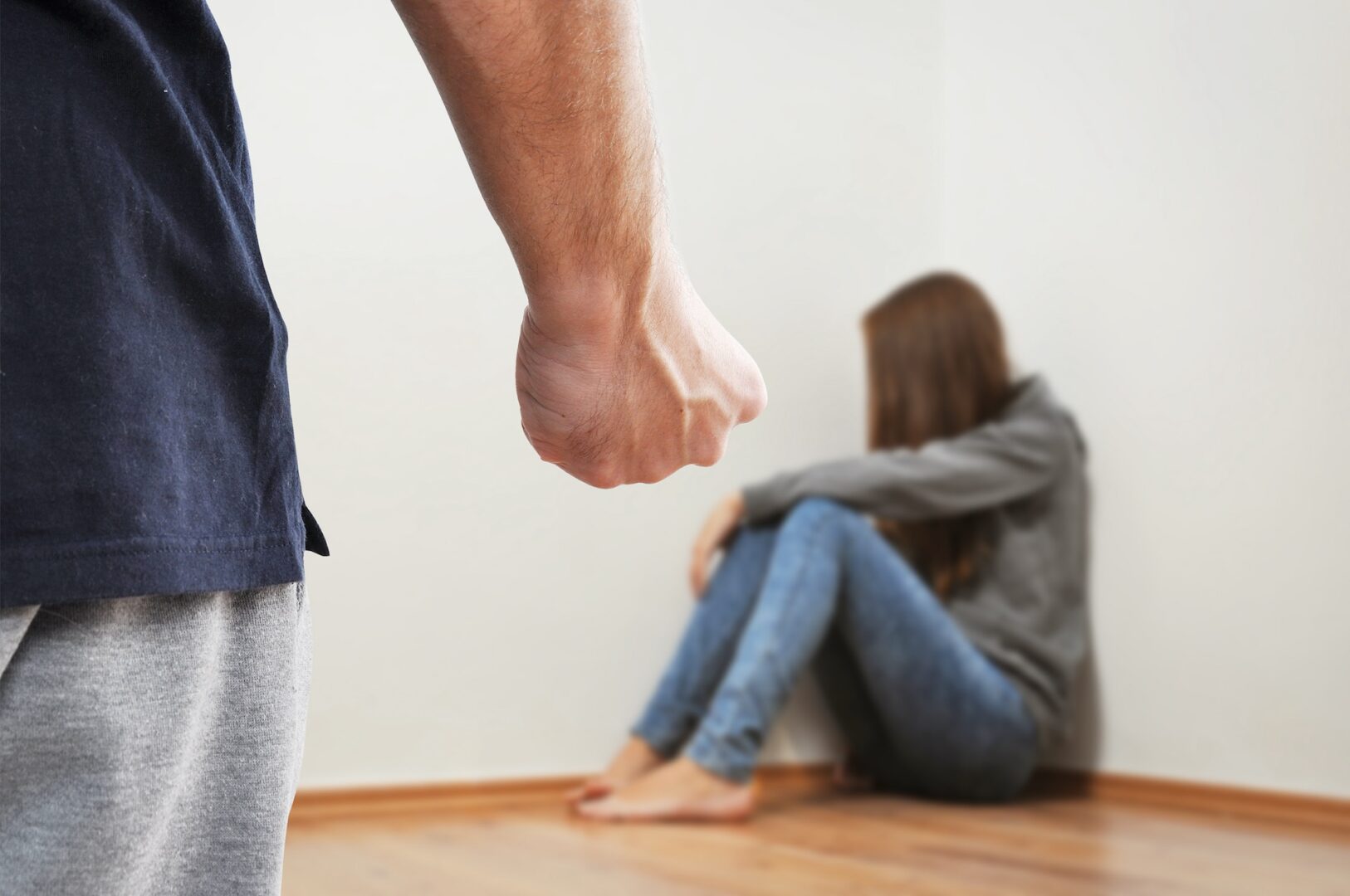 A person with a clenched fist stands in the foreground, while a woman sits on the floor in the corner, looking distressed. She is wearing a gray sweater and jeans. The room has light-colored walls and a wooden floor.