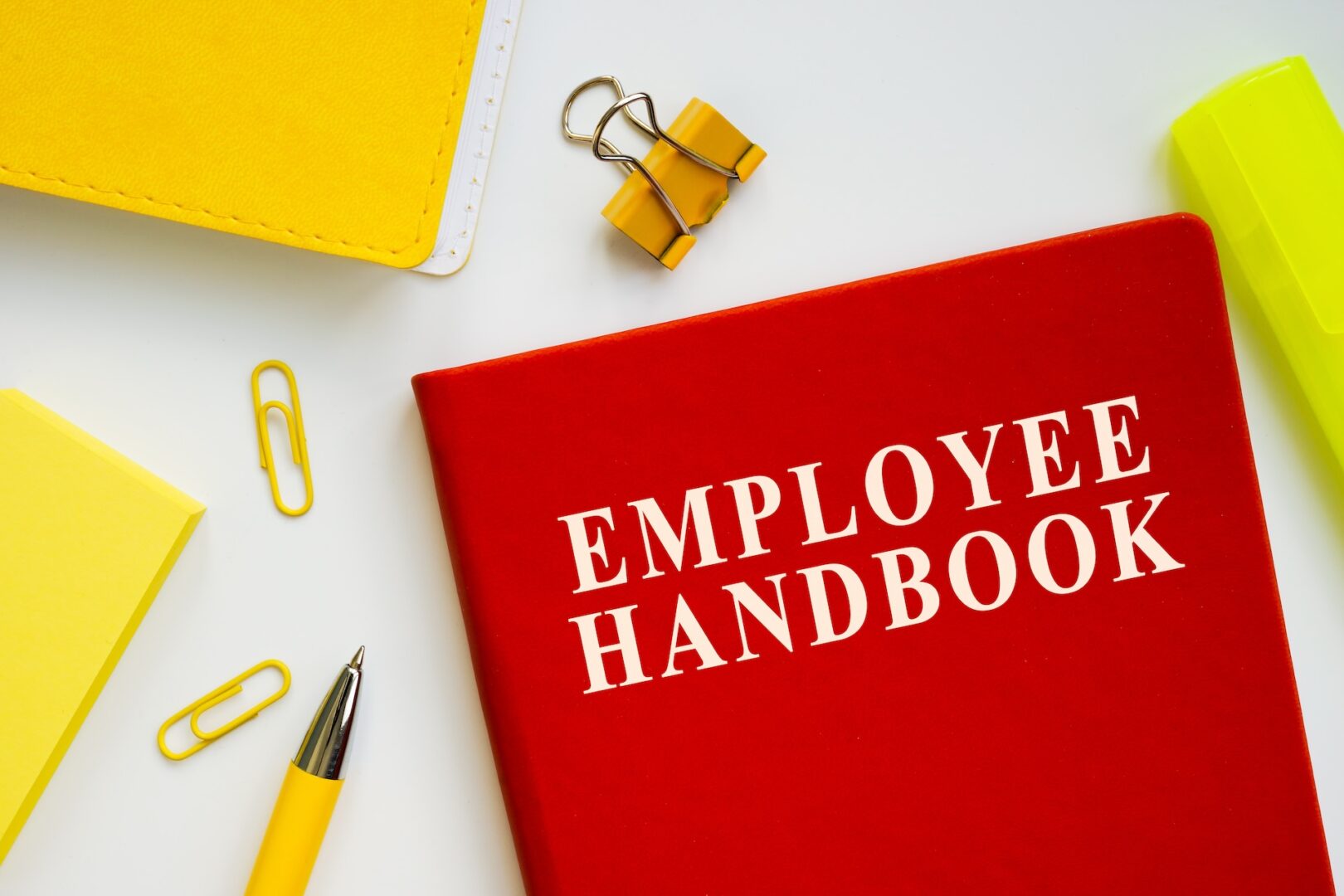 A red book titled "Employee Handbook" is on a white surface, surrounded by a yellow notebook, paper clips, a gold pen, a highlighter, and two yellow binder clips.