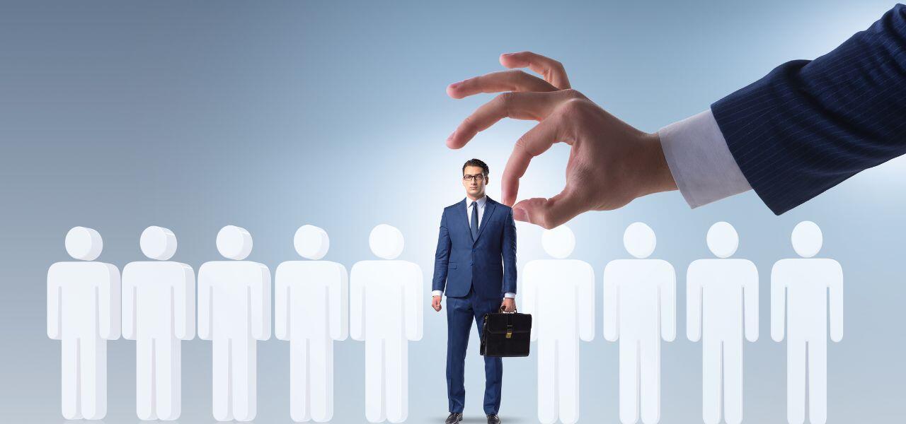 A businessman in a blue suit stands among white silhouettes of people. A large hand in the foreground appears to be picking him out from the group. He holds a briefcase and looks forward confidently.