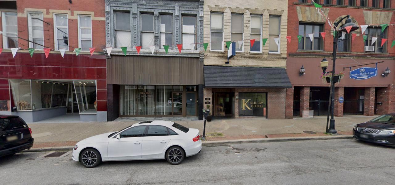 A street view of a row of storefronts featuring colorful flags hung above. A white car is parked in front of a building with a green sign reading "Klein Optics." The buildings have various architectural designs, and the pavement is wet.