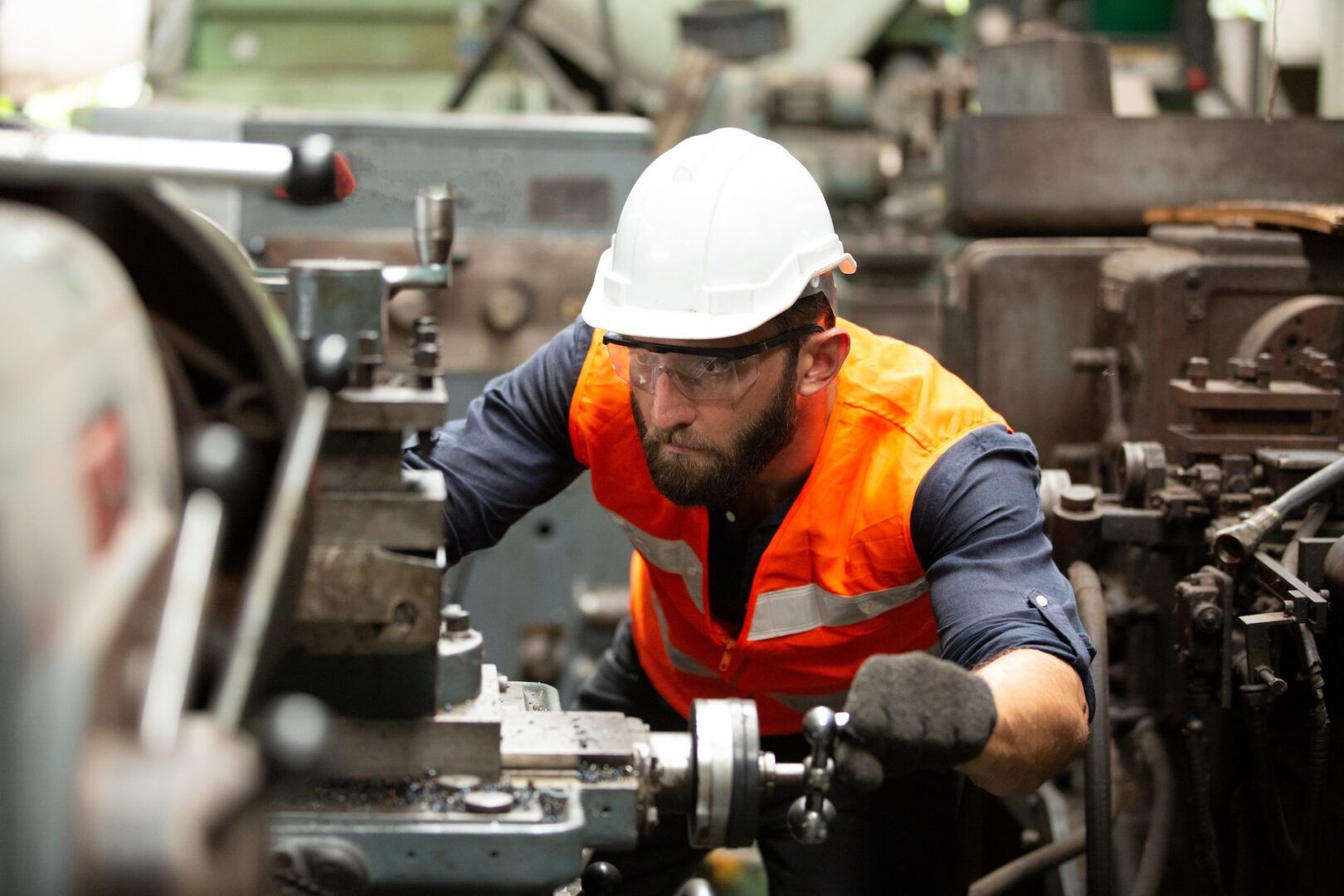A man wearing a white hard hat, safety glasses, and an orange safety vest operates a lathe machine in an industrial setting. He is focused on his work, surrounded by various machinery and equipment. A man wearing a white hard hat, safety glasses, and an orange safety vest operates a lathe machine in an industrial setting. He is focused on his work, surrounded by various machinery and equipment.