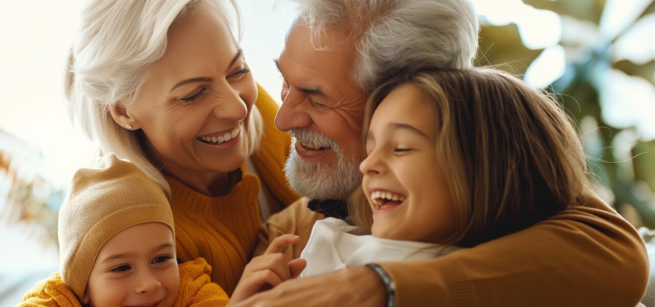 A joyful elderly couple with silver hair embraces two young children, a boy and a girl, as they all laugh together in a warm, sunlit room. The girl is smiling broadly, and the boy is wearing a brown knit hat. The atmosphere is loving and joyful.