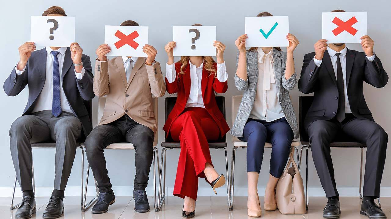 Five people seated in a row holding signs over their faces. Two hold question marks, two hold red X's, and one holds a green check mark. They are dressed in business attire, sitting on chairs against a plain background.