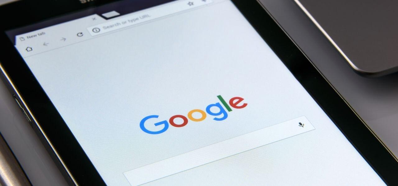 Close-up of a tablet displaying the Google homepage. The screen shows the Google logo above the search bar. The device is resting on a table, with part of its black border visible.