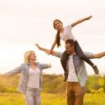 A family of three enjoys a sunny day outdoors. A father carries his daughter on his shoulders while the mother stands nearby. They all have their arms spread wide, surrounded by green grass and trees, with a warm sunset in the background.
