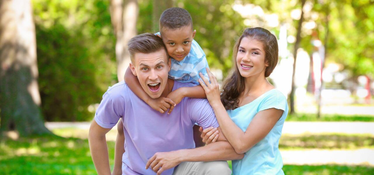 A cheerful family of three poses outdoors in a sunlit park. A man crouches while a young boy sits on his back, smiling. A woman stands beside them, resting her hand on the boy's shoulder. Trees and grass are visible in the background.