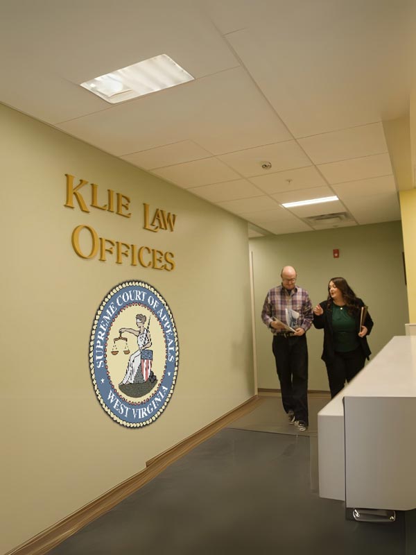 A man and woman walk down a hallway in an office labeled "Klie Law Offices." The wall features the seal of the Supreme Court of Appeals of West Virginia. The ceiling has fluorescent lights, and a white desk is visible.