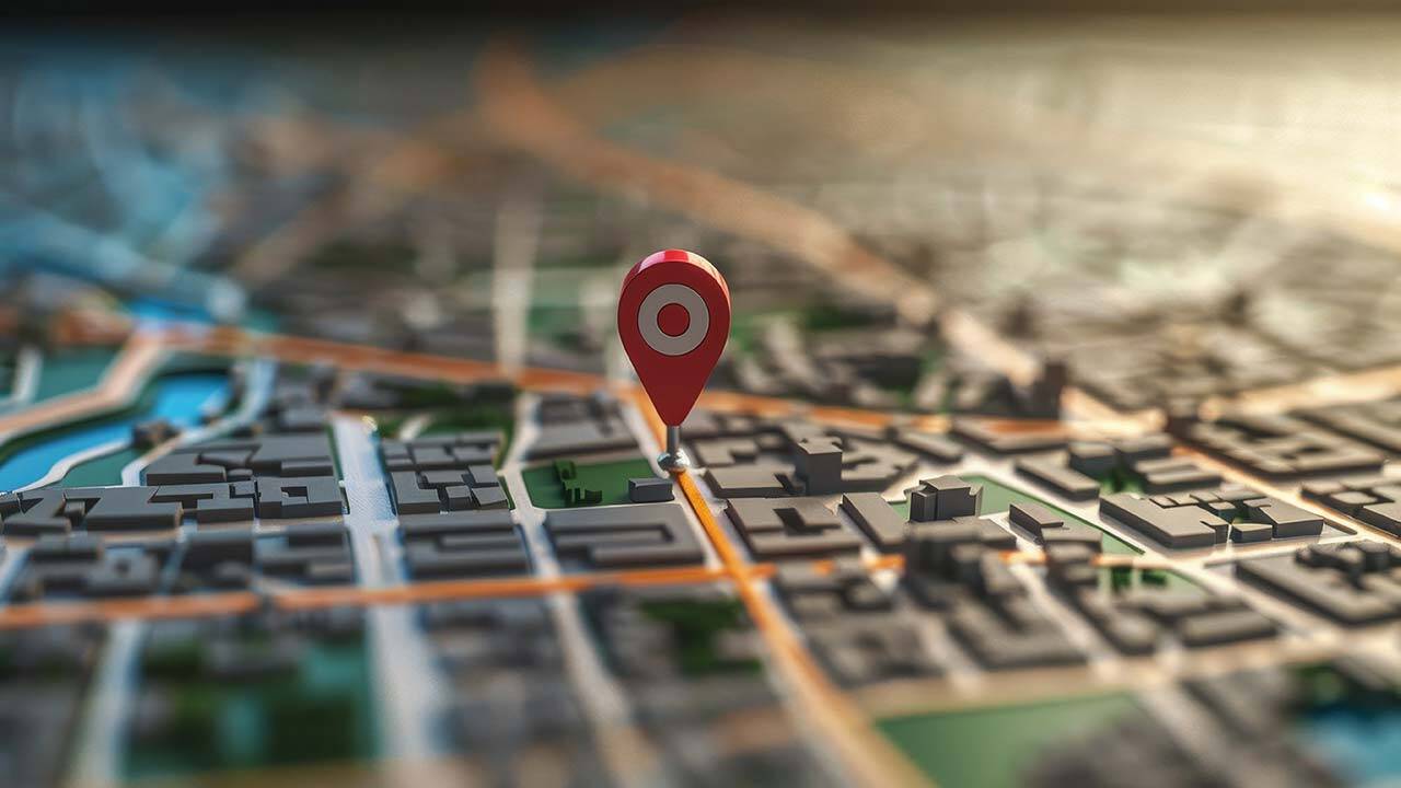 A 3D-rendered map with a red location pin icon hovering above an urban area, highlighting a specific point. Roads and buildings are visible in the background, suggesting a cityscape. The lighting is soft and focus is on the pin.