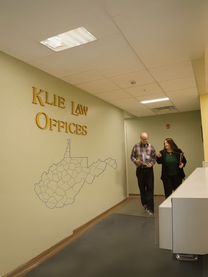 Two people walk down a hallway in Klie Law Offices. The wall displays the firm's name and an outline of West Virginia. They seem to be engaged in conversation, and the area is well-lit with overhead lights.