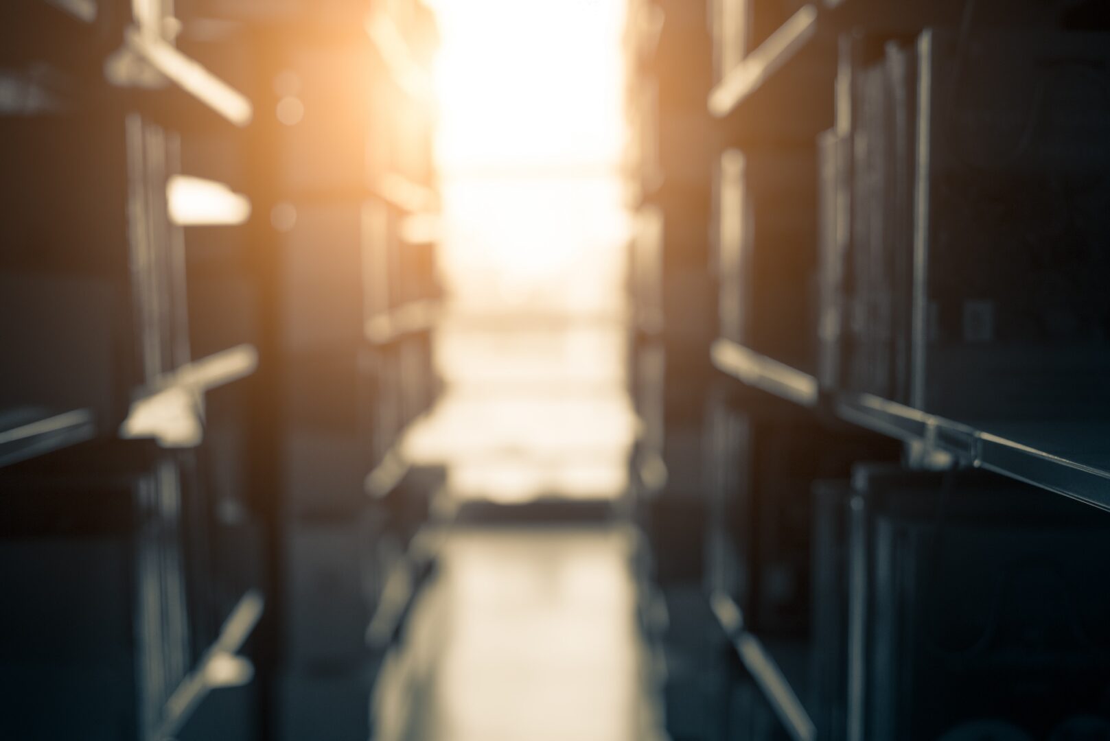 A sunlit library aisle with rows of blurred bookshelves on either side, creating a warm, tranquil atmosphere. The sunlight streams through a window at the end of the aisle, casting a soft glow on the floor.