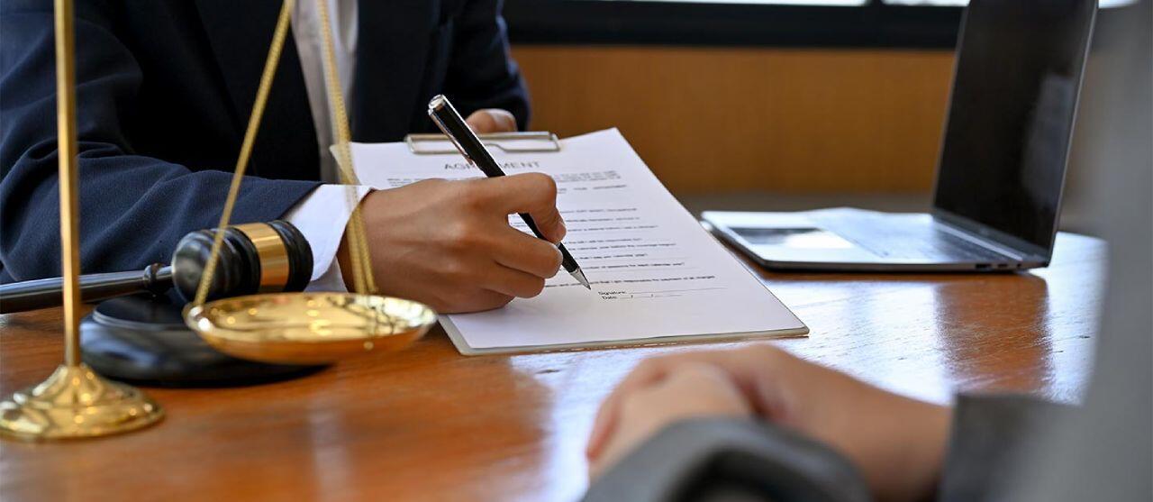 A person signs a document on a clipboard at a desk featuring a gavel, scales, and an open laptop. The setting suggests a legal or professional environment.