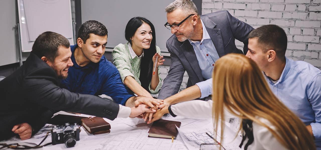 A group of six people in an office environment are gathered around a table, stacking their hands together in a gesture of teamwork and unity. The table is covered with documents and plans. The group appears engaged and collaborative.