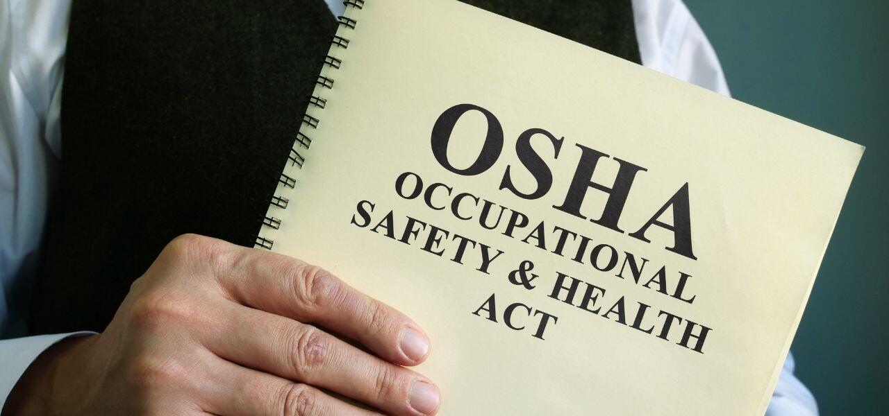 A person holding a spiral-bound booklet titled "OSHA Occupational Safety & Health Act" against a plain background. A person holding a spiral-bound booklet titled "OSHA Occupational Safety & Health Act" against a plain background.