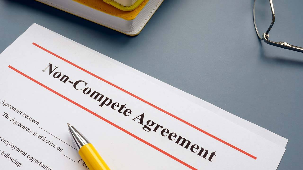 A document titled "Non-Compete Agreement" rests on a table next to a pair of eyeglasses. A yellow pen lies diagonally across the paper, with part of a notebook visible in the background.
