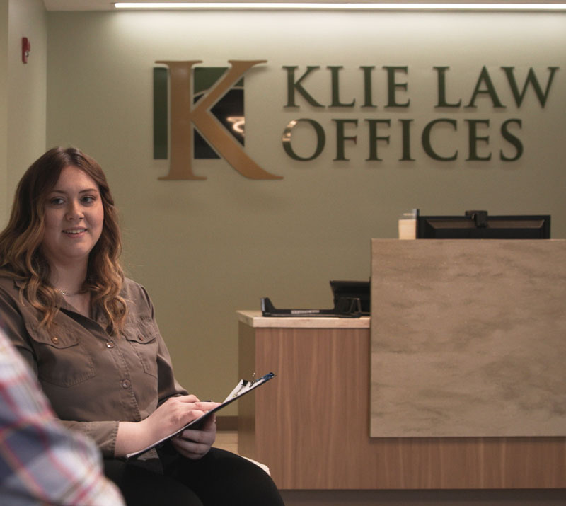 A woman sits in the reception area of Klie Law Offices, holding a clipboard. The office name is displayed on the wall behind her, next to a desk with a computer and a cup. She is smiling and looking towards the camera.