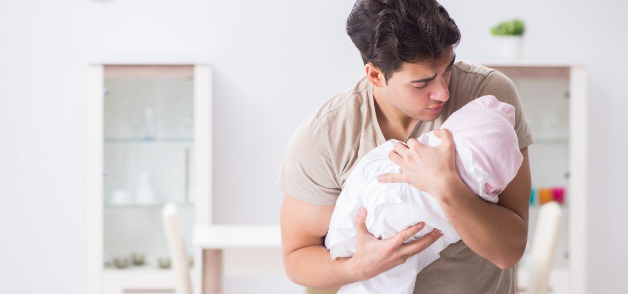 A man gently holds and kisses a baby wrapped in a white blanket with a pink hat. The background features a softly lit room with minimalistic decor, including a cabinet and a potted plant.