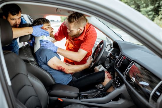 Emergency responders assist an injured man in a car. One responder holds the man's head while the other secures a neck brace. The man appears to have injuries on his arm. The car is parked, and the responders are focused on providing aid.