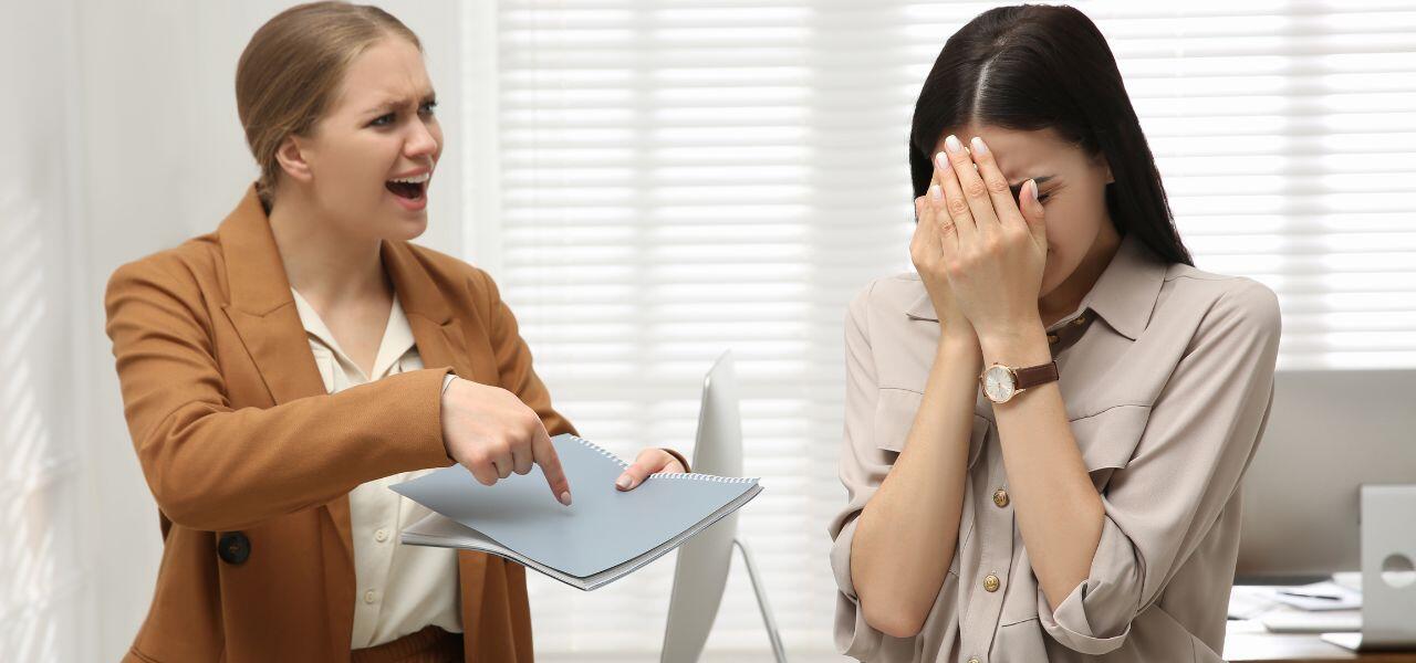 A woman in a brown blazer angrily points at a document while speaking to another woman who is covering her face in distress. They are in a bright office setting.