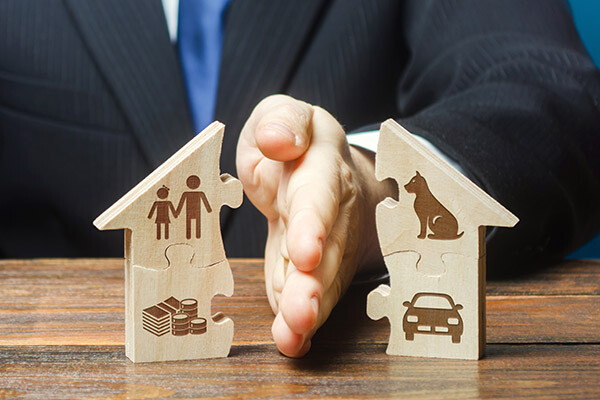 A person in a suit using their hand to separate two wooden house-shaped puzzle pieces. One piece has icons of a family and money, while the other shows a car and a dog, symbolizing division of assets.