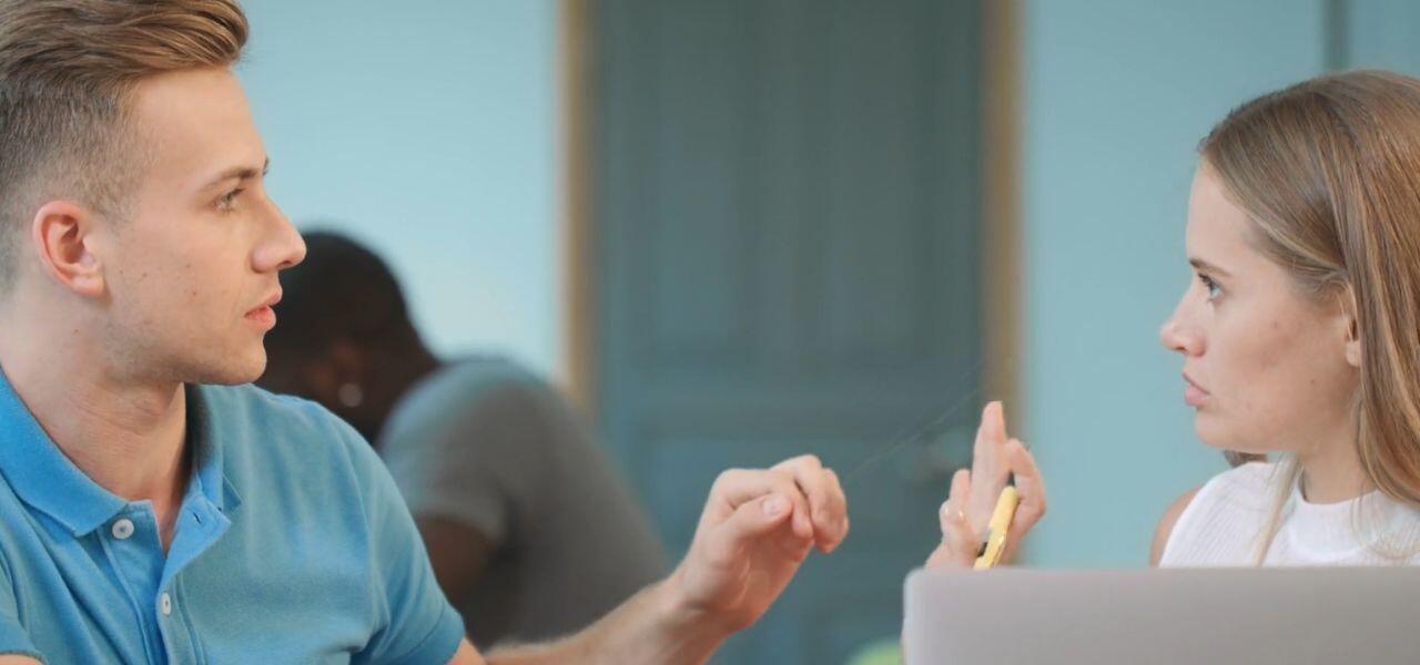 A man and woman are seated in an office setting, engaged in a discussion. The man is gesturing with his hands, while the woman holds a pen and listens intently. A laptop is partially visible in front of them, and another person is in the background.