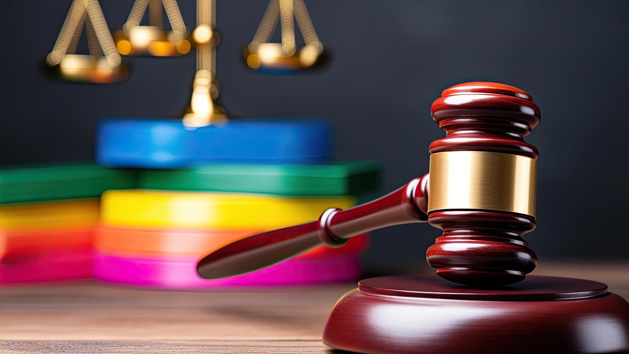 A wooden judge's gavel rests on its sound block in the foreground. Behind it, scales of justice are visible with a blurred stack of colorful circular objects, suggesting a concept of law and order alongside diversity.