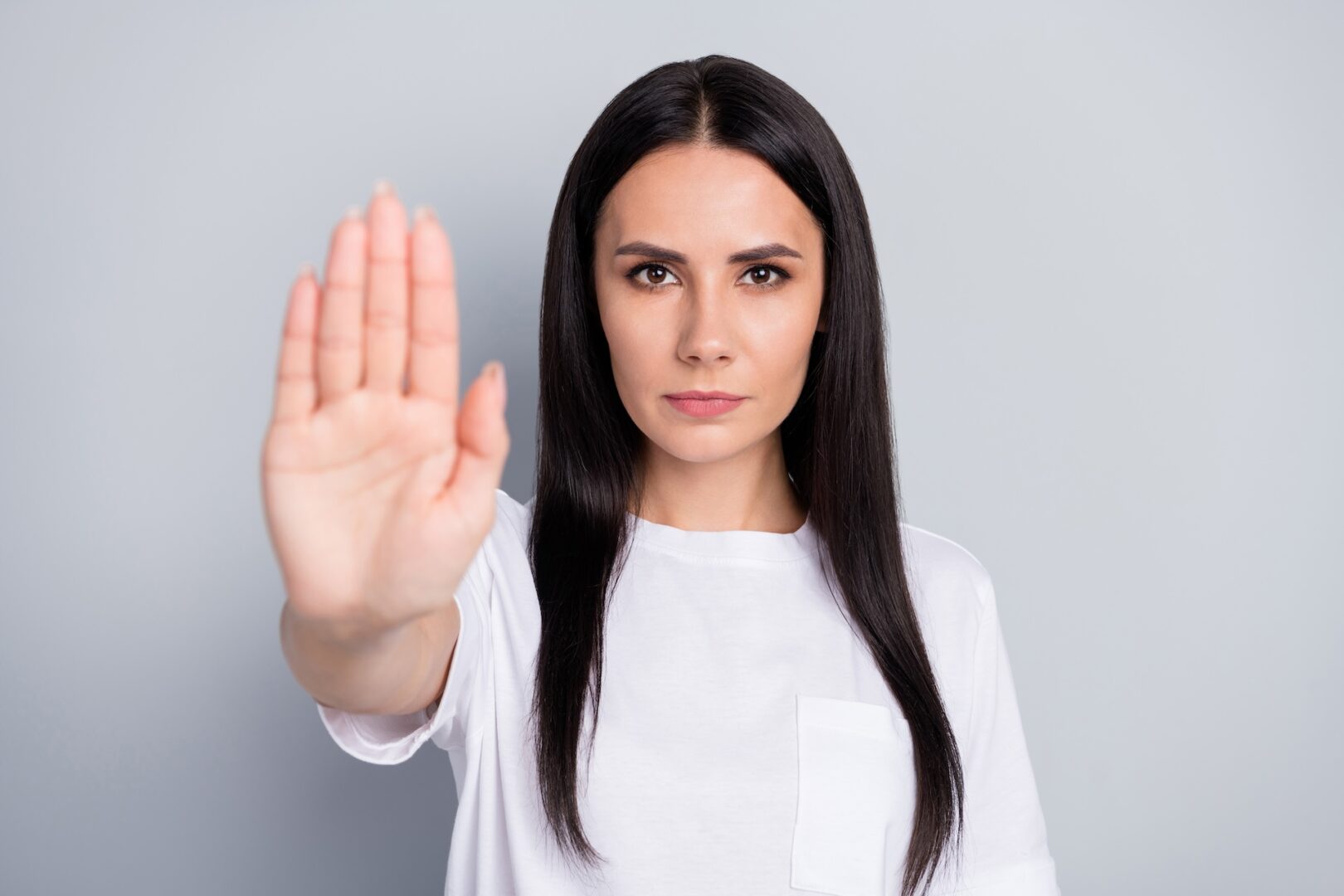 A woman with long dark hair wearing a white shirt holds her hand out in front of her, palm facing the camera, as if signaling "stop." She stands against a plain gray background.