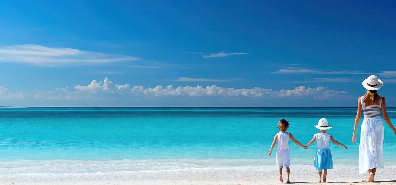 A woman and two children in white and light blue clothing and hats stand hand in hand facing the turquoise ocean on a sandy beach under a clear blue sky.