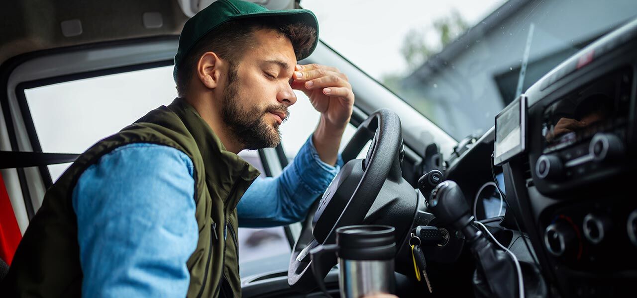 A man sitting in the driver's seat of a vehicle looks tired, resting his forehead on his hand. He's wearing a cap and holding a travel mug. The dashboard and steering wheel are visible in the foreground.