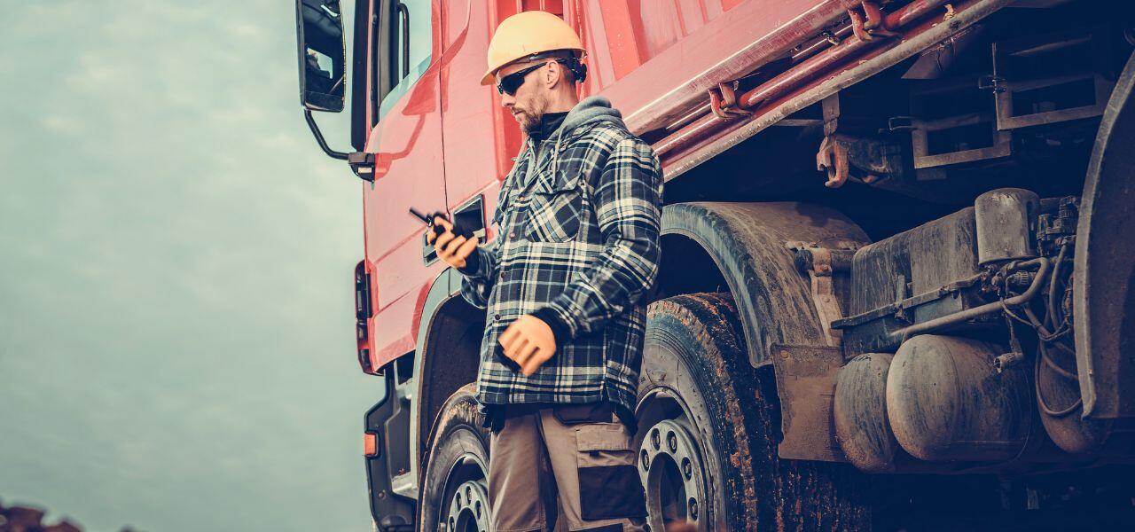 A construction worker wearing a hard hat and plaid jacket stands beside a large red truck, holding a walkie-talkie. The scene suggests a worksite environment with a focus on communication and safety.