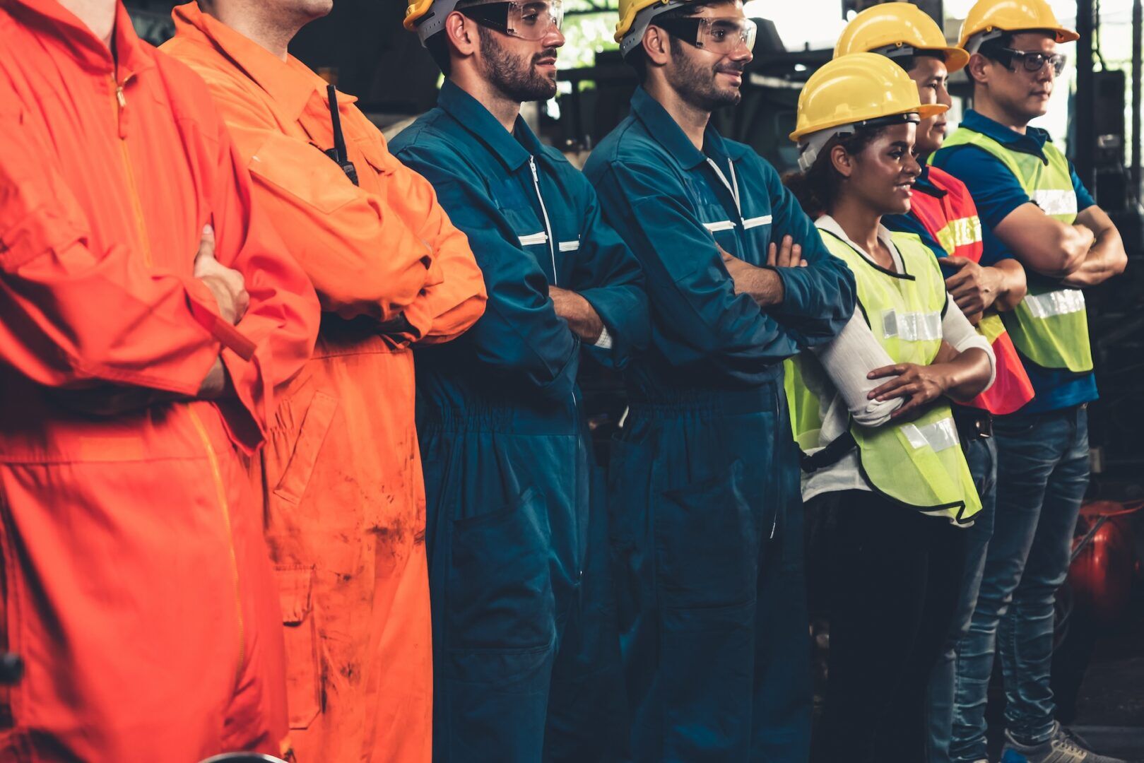 A group of industrial workers stands in a line wearing protective gear. Some are in orange coveralls, others in blue, while a woman wears a neon safety vest and yellow hard hat. They appear focused and attentive.