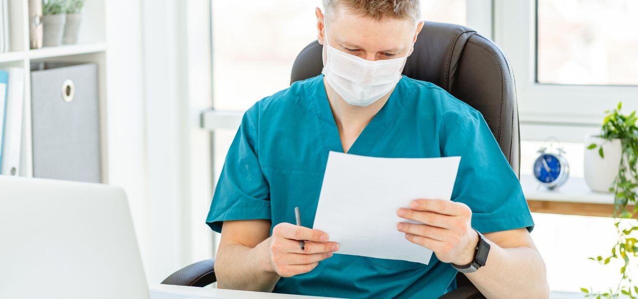 A healthcare professional wearing a surgical mask and blue scrubs sits at a desk, reviewing a document. A laptop is open beside them, and a clock and potted plant are visible in the background.