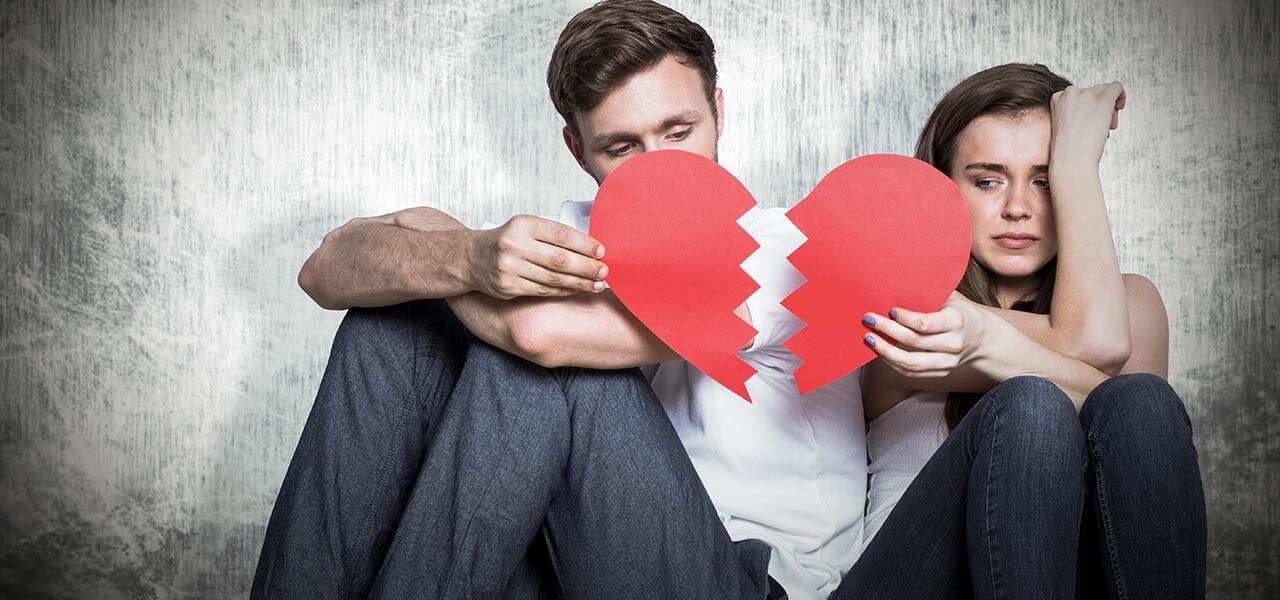 A man and woman sit on the floor looking sad, holding two pieces of a torn red paper heart. The backdrop is a textured grey wall, and both appear pensive and distant.