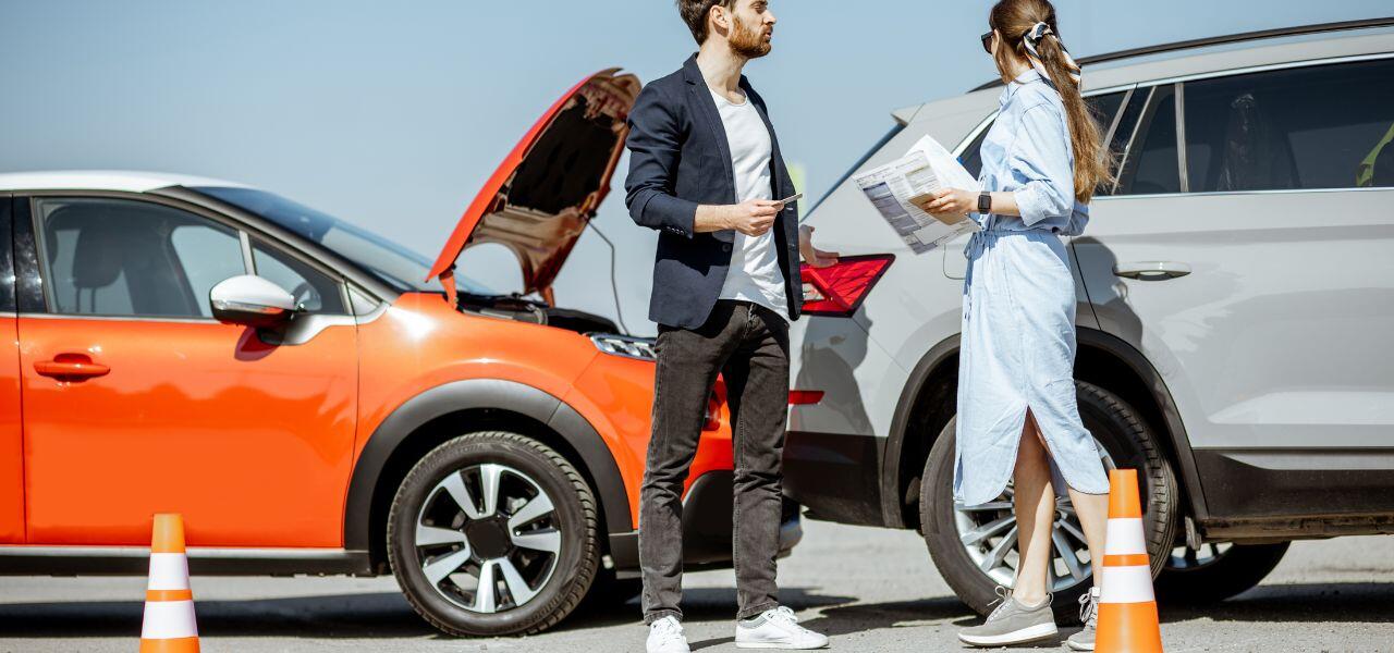 A man and woman stand between two cars that have been in a minor collision. The red car's hood is open. They're holding documents and discussing the situation. Two traffic cones are placed in front of the vehicles.