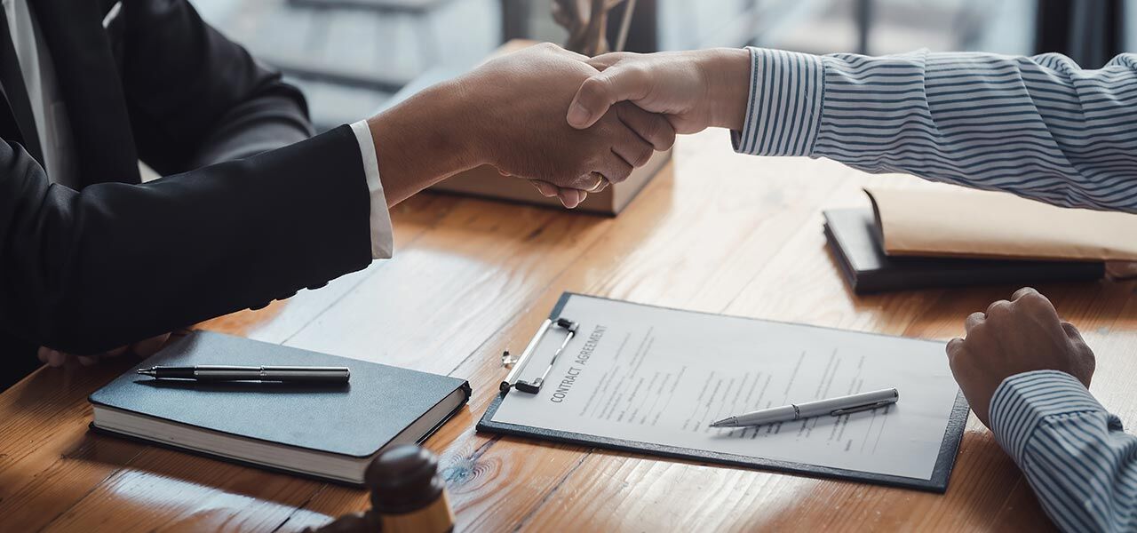 Two people shake hands over a wooden table. Documents and pens are on the table, including a contract agreement on a clipboard. Both are wearing business attire, suggesting a professional meeting or agreement.